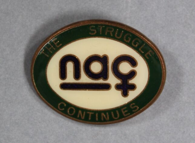 An oval shaped badge with 'NAC' in the centre. The C is made to look like the 'woman' symbol. Around the outside of the badge it reads, "The Struggle Continues"