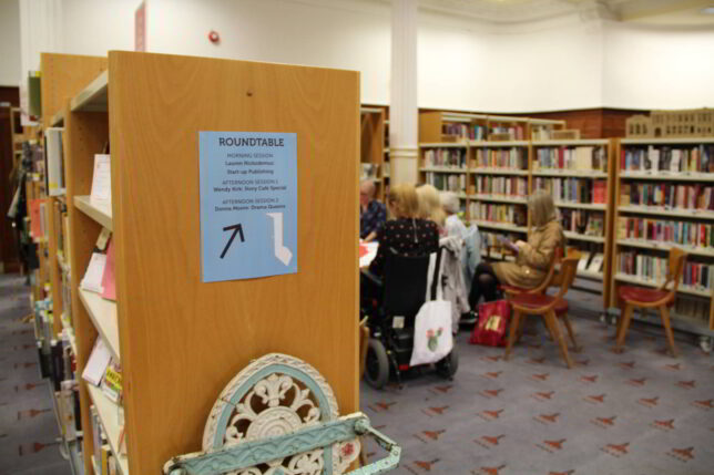 The Library space with a poster stuck to the end of one of the bookshelves. It indicates the workshops taking place in the space. In the background you can make out some women gathered around a table.