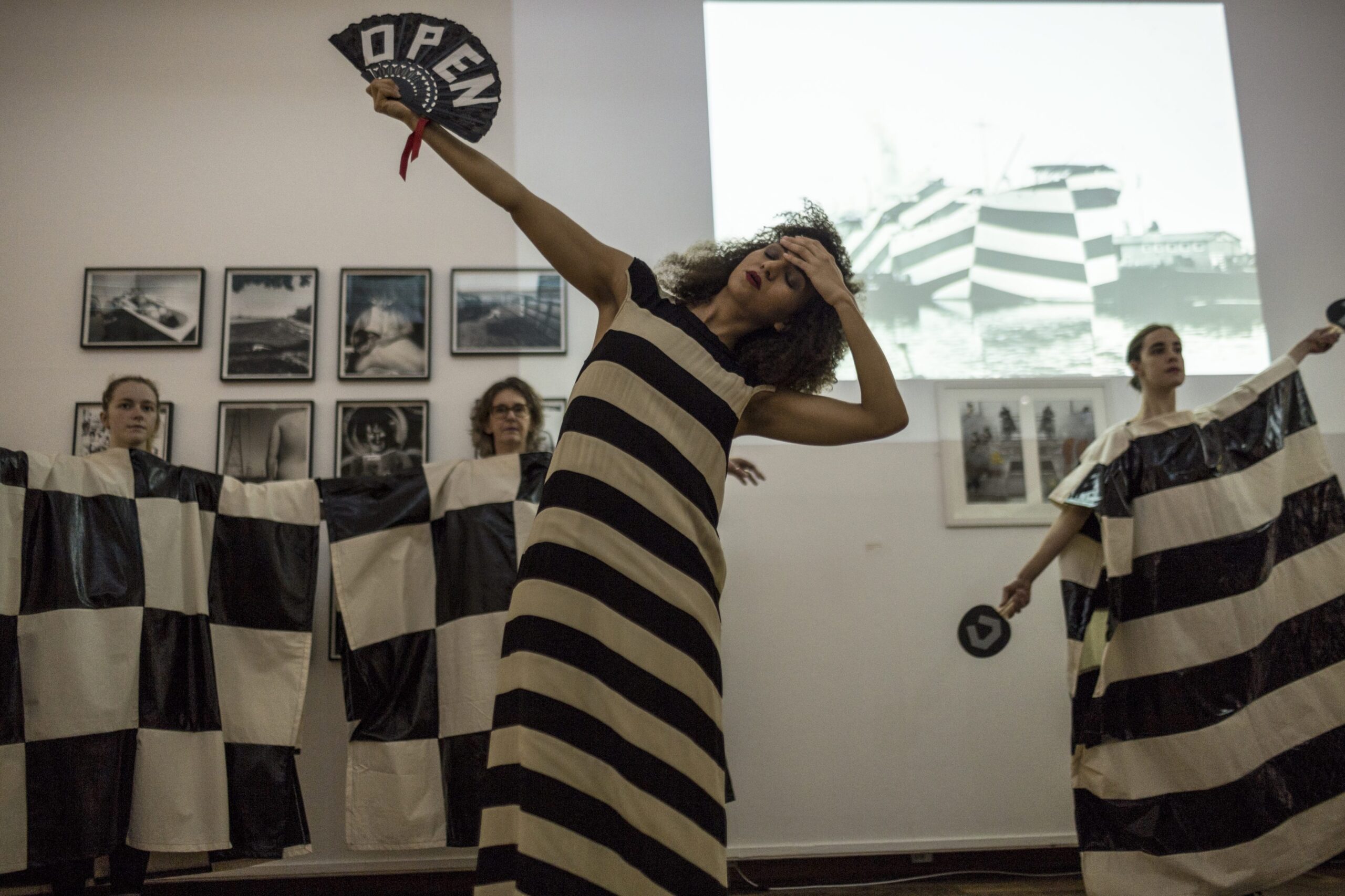 Image of Linder, Love Imposes Impossible Tasks, Performance - a person in a black and white striped dress holds up a fan saying 'OPEN' while holding the back of their other hand to their forehead and swooning. 3 people in black and white geometric costumes are lined up in the background.