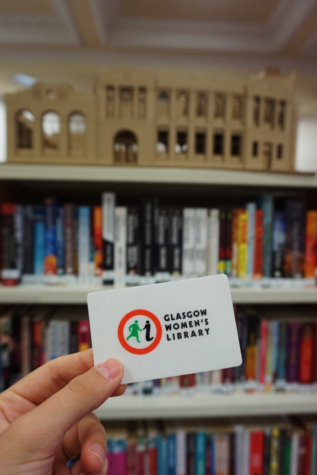 A hand holds up a GWL Borrower Card bearing the GWL logo, a red circle with a green figure of a woman and an 'i' for information. There are bookshelves and an architect's model of the building behind.