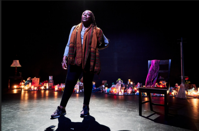 A black woman standing on stage with a scarf around her shoulders. Lights are lit on the floor behind her and a chair sits to the right of her.