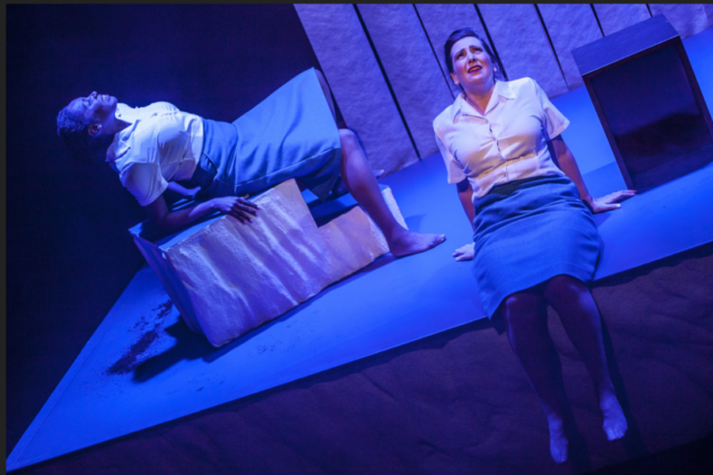 A shot from a performance. Two women sit on the stage and are lit with a blue tint.