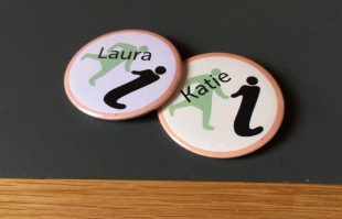 Two Glasgow Women's Library design badges laying on a worktop. The badges have the GWL logo of a woman holding a 'i' or info symbol. On the badges are the names Laura and Katie.