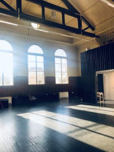 Three very large 1900s style windows flood the room with light, the black floor with shadows and bright sunlight sections.