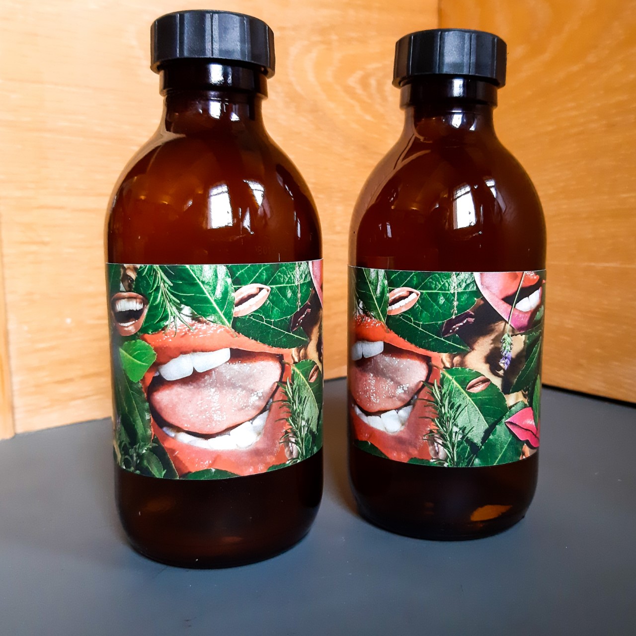 2 Brown bottles with black lids, with Linder's design on the label. The design is of open mouths with pink lips, with greenery and leaves surrounding it.