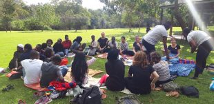 A group of about 20 people sitting in a circle on the grass in a park on a sunny afternoon