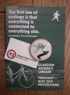 A badge with the name "Lily" on it, sitting next to a GWL library card on the GWL programme of events from Februay to May 2019. There is a quote from Nan Shpherd on the programme: The first law of ecology is tat everything is connected to everything else".