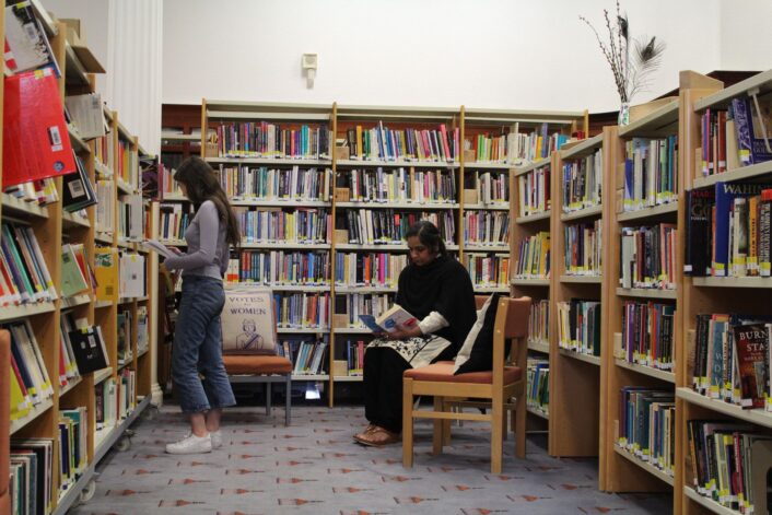 Amongs the GWL shelves, a woman sits reading a book, wile another stands browsing the shelves.