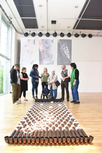 A group of adults stand together in a gallery, there is an artwork on the floor similar to a raft made from black rain pipes tied together with pink fabric.