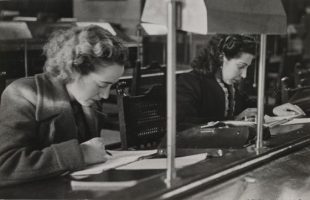 Black and white imagephoto of two women leaning over documents on a desk, pen in hand. Looks like the photo was taken in the mid 20th century.