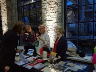 Some of the Community Curators discuss their work with a visitor, beside a table displaying items from the GWL collection including photographs, postcards, badges and a fluorescent yellow, orange and brown curly wig. 