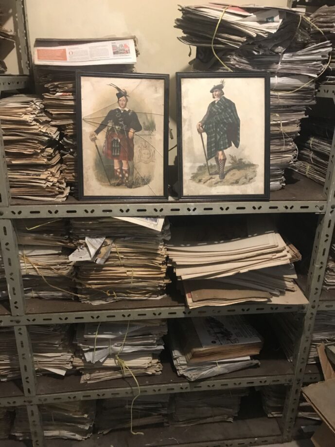 Framed prints of Scotsmen in the stores at McMillan Library. The prints, which are old-fashioned fading prints of men in kilts, are propped up on metal shelves full of papers.