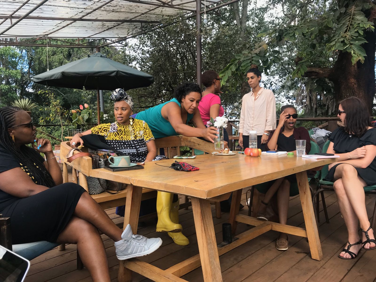 Feminist leaders assembling for gathering, Feminist Governance: A Nairobi perspective. Seven women, mostly women of colour, sit and stand talking together on a balcony with trees beyond.