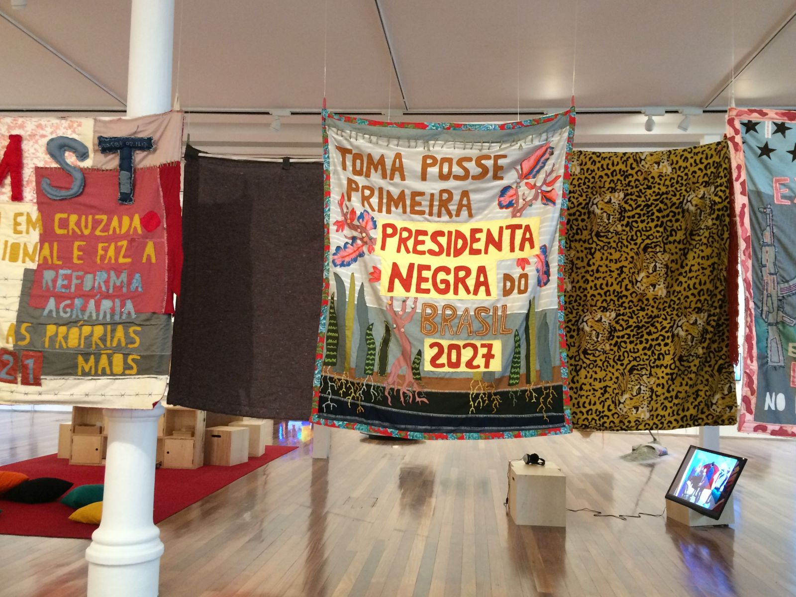 Artworks by Randolpho Lamanier, 2018. Several colourful banners hang in an high-ceilinged room. The central banner, featuring stylised plants in green, pink and blue, reads 'Toma posse primeira Presidenta Negra do Brasil 2027' ('First Black President of Brazil takes office 2027')