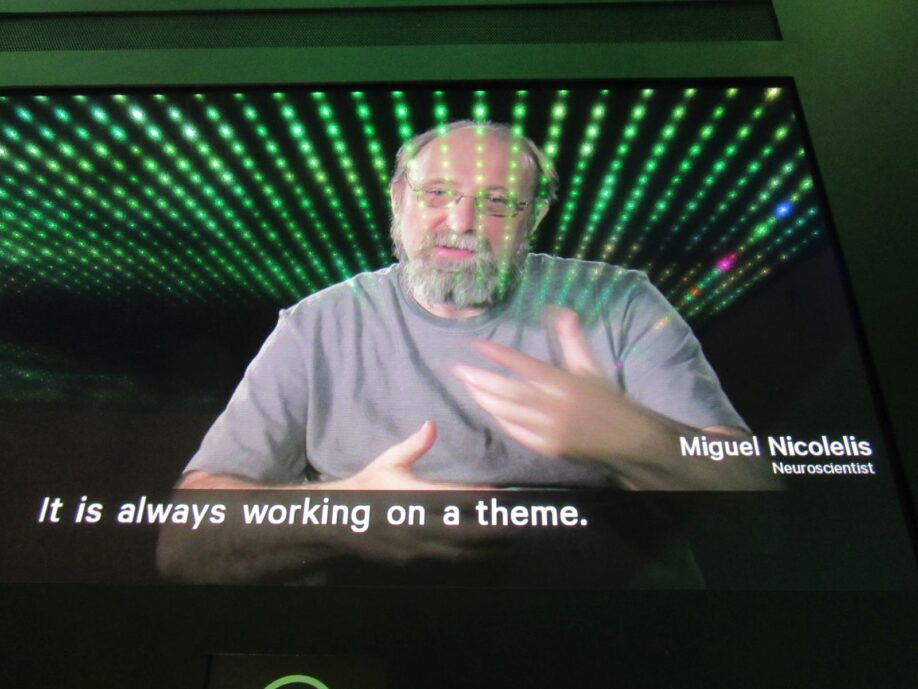 Photo of a video screen featuring a balding, grey-bearded white man gesturing as he speaks. The subtitles on the screen read 'It is always working on a theme'.