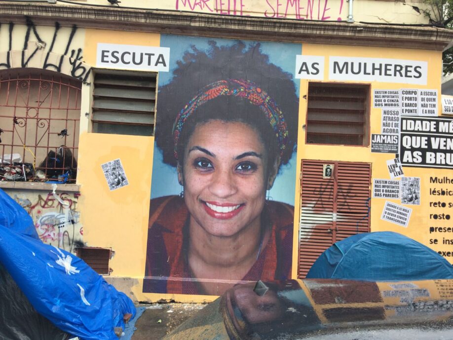 Street artwork memorialising murdered politician, feminist, lesbian activist Marielle Franco. A large photo of Marielle Franco with a vivid smile has been pasted on a wall; text aound the photo reads 'Escuta as mulheres' ('Listen to women').