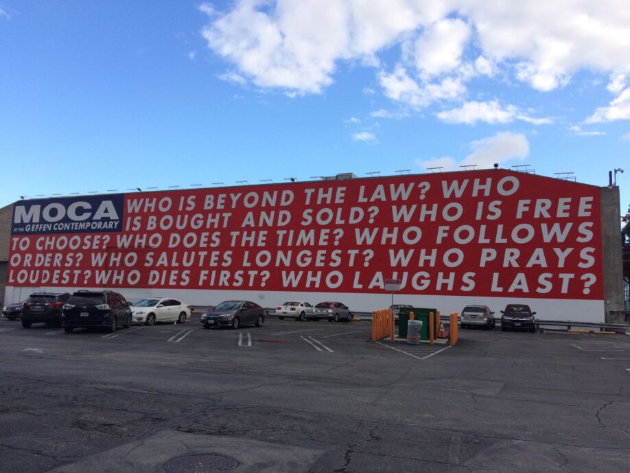 Barbara Kruger (reinstalled) wall work, Untitled (Questions) (1990/2018). On a large building, in white, bold, sans serif capitals on a red background, the 9 questions are: ‘Who is beyond the law? Who is bought and sold? Who is free to choose? Who does the time? Who follows orders? Who salutes the longest? Who prays loudest? Who laughs last?'