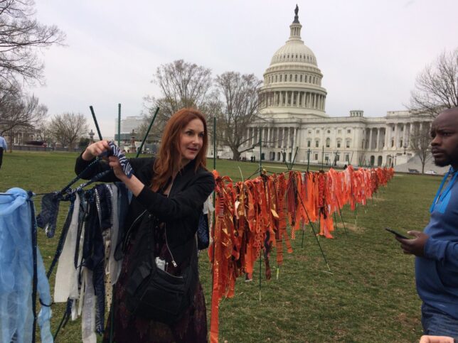 Art activist adding pieces of fabric to a work outside the White House, Washington, representing the 13 thousand children killed by guns in the period between the murder of children at Sandy Hook and March 2019. A long line of scraps of fabric, mostly in shades of red, stretches into the distance, where the White House can be seen.