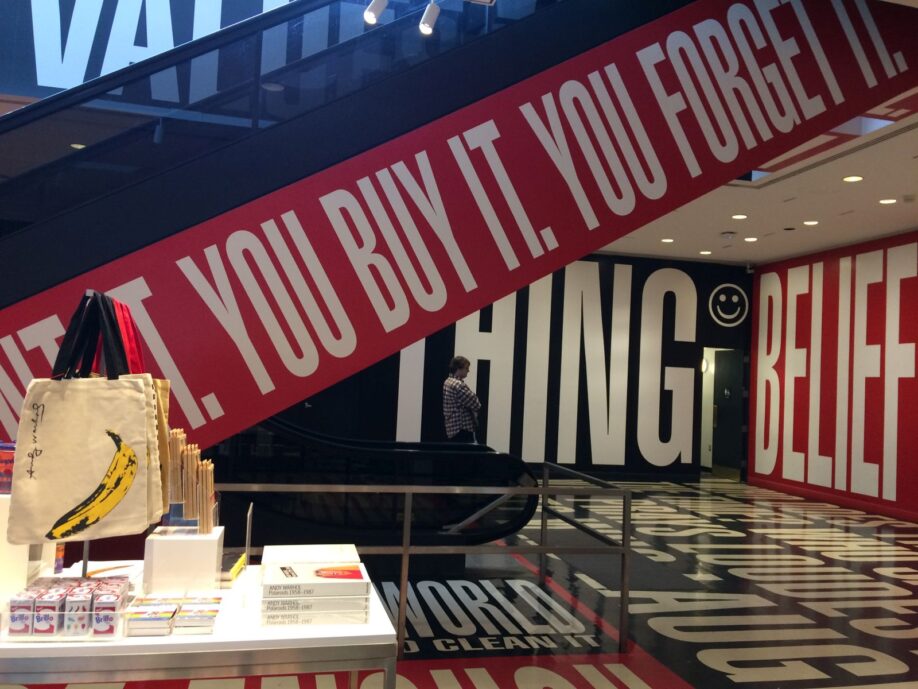 Detail of Barbara Kruger artwork Belief and Doubt, 2012, (You want it, You Buy it, You forget it) next to museum shop display. 'You want it, You Buy it, You forget it' runs in bold, white capitals on a red background up an escalator; sections of further slogans can be seen on the walls beyond.