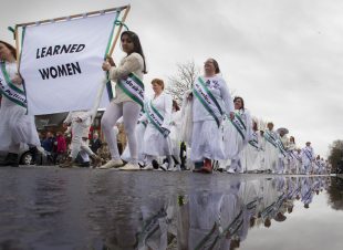 Women dressed in white with purple and green sashes process down a road.