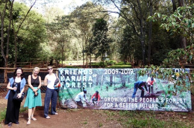 Three smiling women standing beside a large banner stating 'Friends of Karura Forest 2009-2019: Growing the people's forest for a better future' with the forest nursery behind a fence.