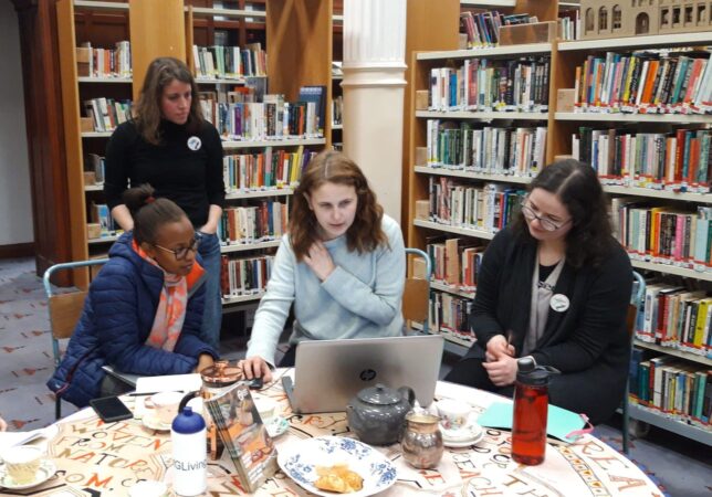 Four women working around a laptop at a round table filled with tea cups, buns and GWL programmes, with stacks of bookshelves behind.