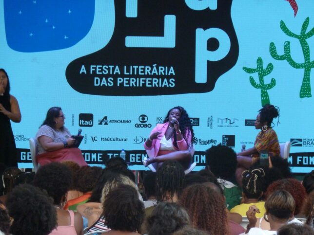 Flávia Oliveira and Roberta Estrela D'Alva, Akua Naru and Preta Rara interviewed by Jéssica Balbino - they are sitting on a low stage with the FLUP festival logo projected behind them