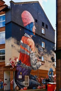 Mural on gable end of a block of flats, showing a woman with blue hair, a pink hat and an orange high vis vest, and flowers growing around her.