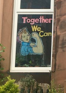 Paper cut out stuck to the inside of a window of the words "Together we can" and a drawing of Rosie the Riveter, a woman in blue overalls and a red head scarf flexing her arm muscle (from the 1940's poster)