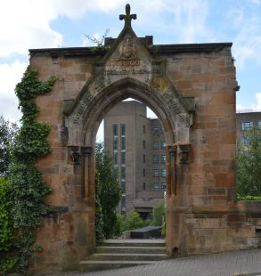 Sandstone archway with a cross on the top, with ivy growing up on side. 