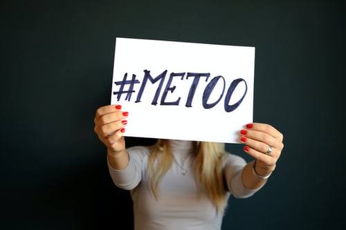 Picture of a woman holding up a sign in front of her face with the hashtag me too written on it.