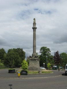 The monument is a tall column on a pedestal both made of pale stone. It stands in the middle of a roundabout