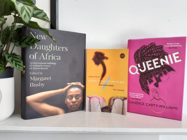 Three books standing on a shelf with a plant. The books are New Daughters of Africa, edited by Margaret Busby, which has a black cover with a photo of a black woman looking directly at the reader, her hands clasped behind her head; Where the Wild Ladies Are, by Matsuda Aoko, a yellow cover with a dark abstract shape in the top half, and an illustration of the knees of two women in skirts sitting beside each other; Queenie, by Candace Carty-Williams, a deep pink cover with the head of a black woman in profile, her face not visible, and long braids pulled up into a bun.