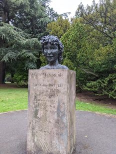 Bust of a woman sitting on a pedestal of stone engraved with the name "Sheila McKechnie". The monument appears to be in a park with tress in the background.