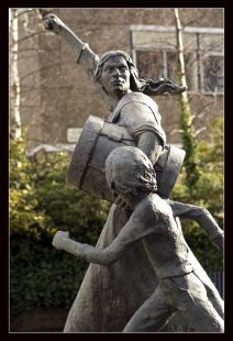 Statue of two people marching: a determined looking young woman holding a drum which she is beating with a clenched fist, and a child walking alongside her and looking up to her, whose face we cannot see