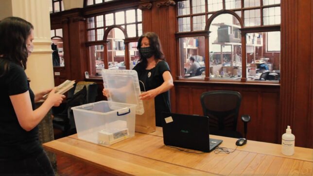 Screenshot from GWL Covid-19 Safety Procedures video - our librarian opens a plastic box for a visitor to place a returned book into. They are both wearing facemasks.