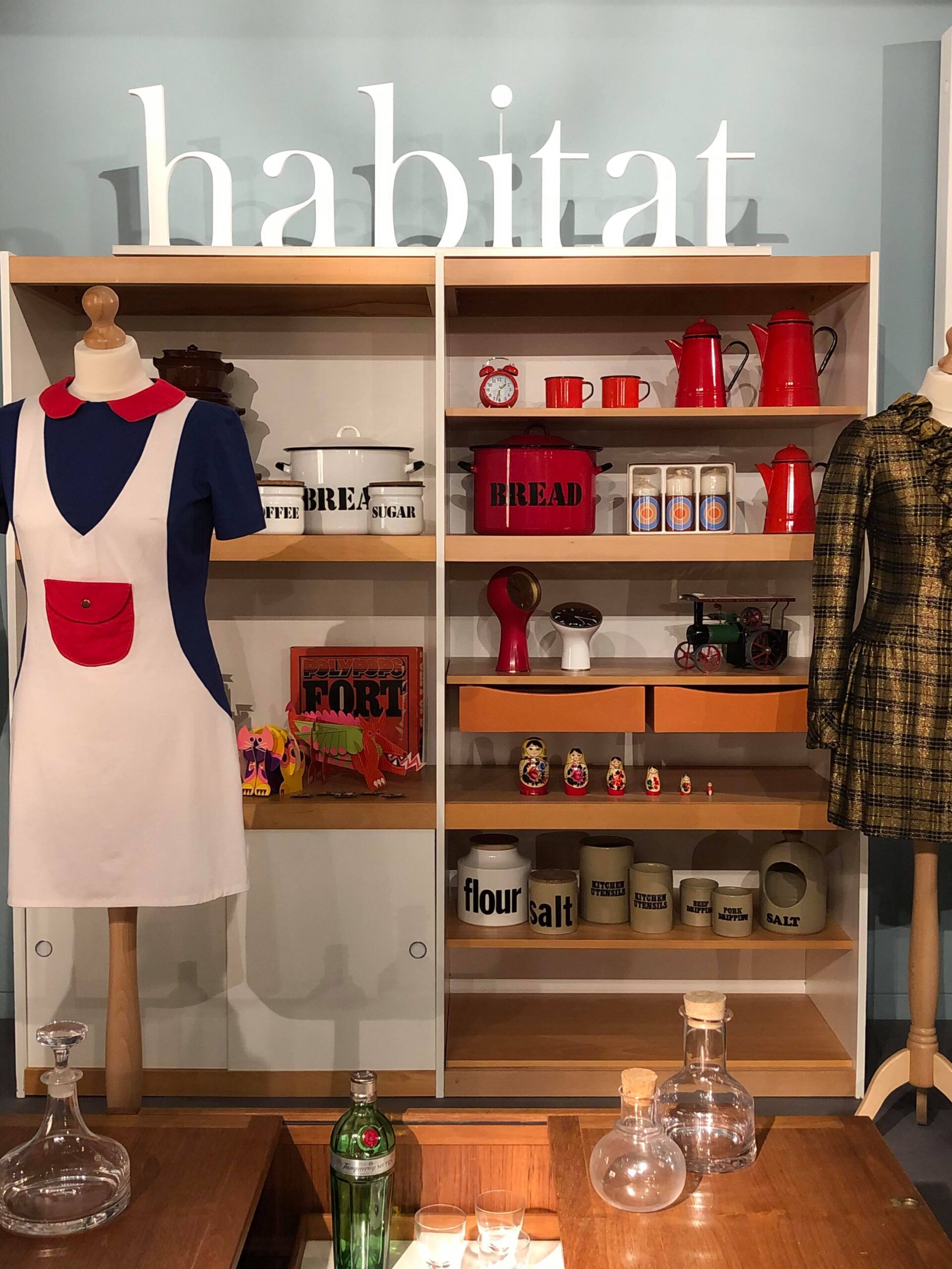 This photo shows a pale blue background wall in front of which a large shelving unit stands. On top of the uni is a large white 'habitat' sign and the shelves hold several household habitat designs including bread bins, alarm clocks, and coffee pots. In front of the shelves are two mannequins, one wearing a blue red and white pinafore style minidress, and one wearing a shiny gold and brown checked lame party dress.