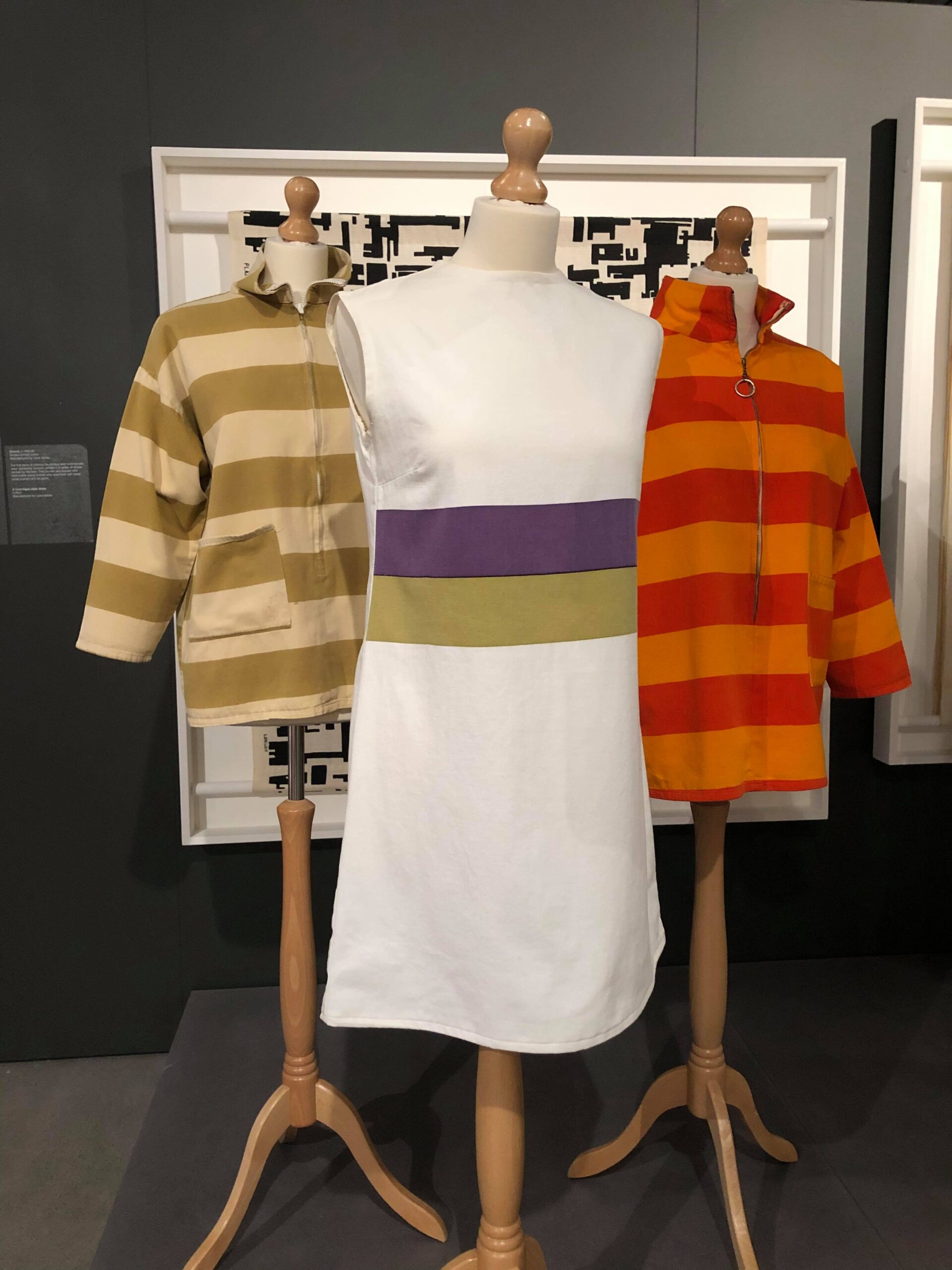 This photo shows a black background wall, with a large white square frame hanging on it. In the frame is a sample of a black and white patterned fabric. In the foreground are three mannequins. Two of them wear striped canvas gardening smocks, on the left is a beige and green one, on the right a bright orange and red. The front mannequin wears a white basic sleeveless minidress with two thick stripes across the waist, one in purple and one in green.