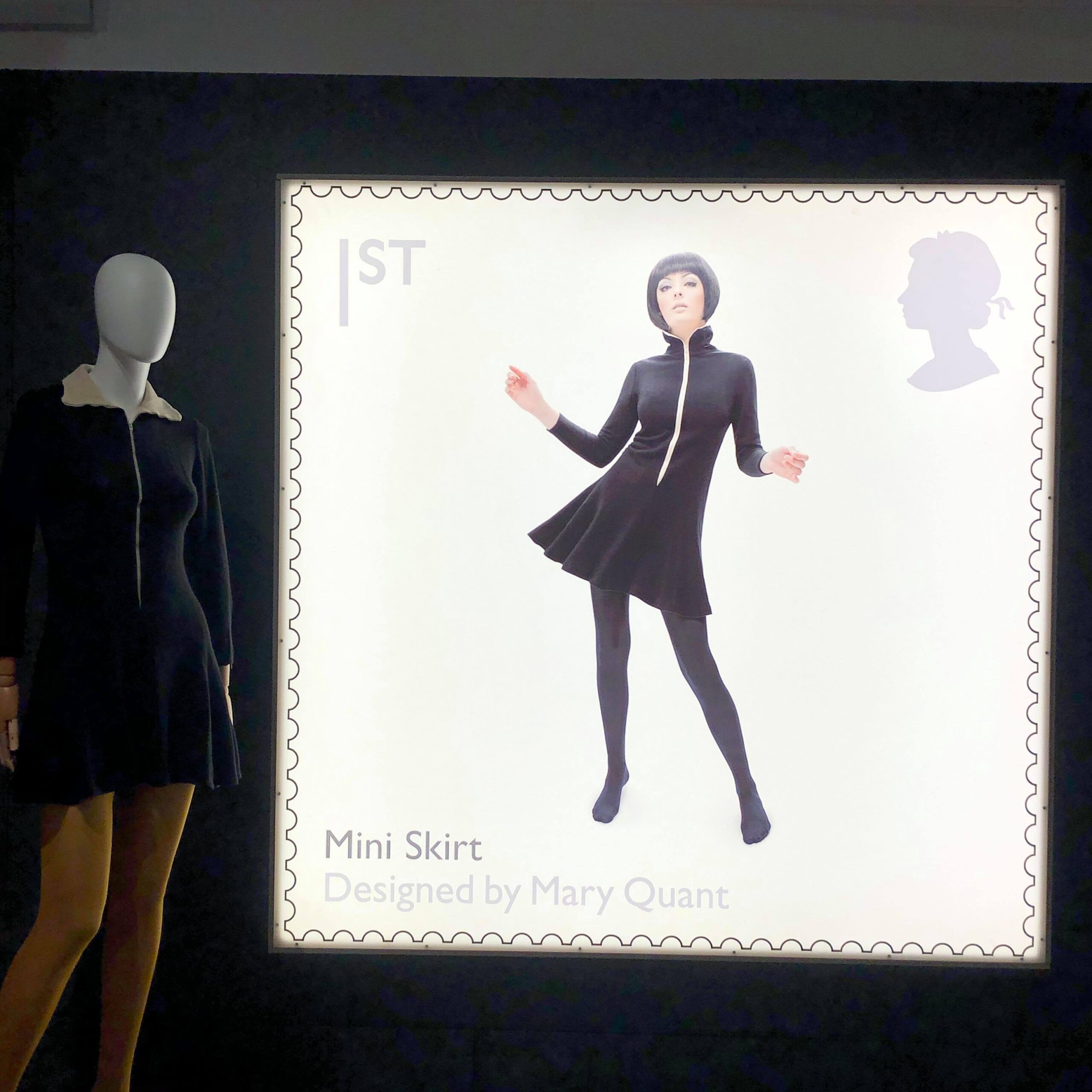 This photo shows a dark background wall with a large illuminated square image of a first class stamp. Mary Quant herself models one of her early mini dress designs - an all black, long-sleeved garment with a white zip down the front. She wears black tights and no shoes. To the left of the stamp is the same mini dress on a white mannequin over yellow tights. Here the collar is turned down and we see the lining is white