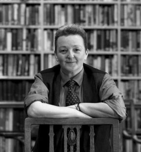 Portrait of Dr Noir in black and white. They are leaning on the back of a chair in front of a bookcase.