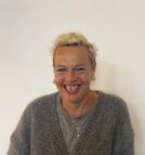 Adele stands against a white wall. Adele has cropped blonde hair and a big smile. She is wearing a grey wooden cardigan over a grey t-shirt