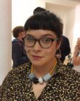 Naomi is wearing glasses, a leoprad print shirt and large beads around her neck. Naomi has dark hair tied up with a fringe