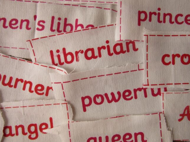 Detail of a pile of white cloth scraps that have different derogatory everyday words for women and feminists printed on them in red.