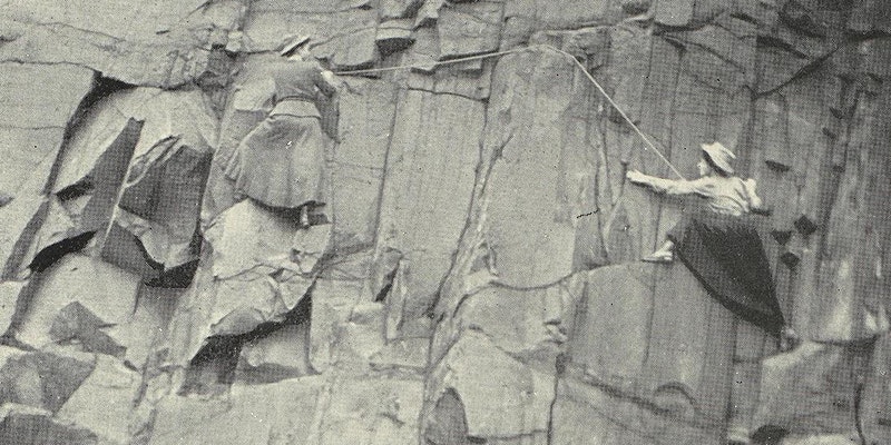 Two women wearing skirts are scaling a cliff in a black and white photo.