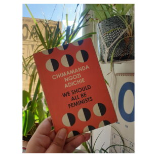 a hand holds the small book "We Should all Be Feminists" at a window with plants and blue skies in the background