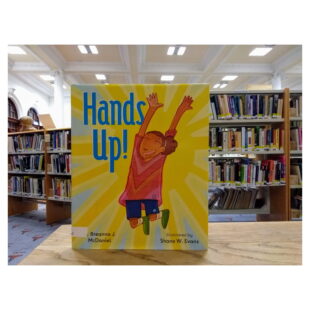 a yellow book cover with the words "Hands up!" written in blue next to an illustration of a child jumping up sits in the library in front of bookshelves