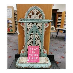 a bright pink book sits on a wrought iron umbrella stand. The book is pink with bold upper case white text which reads "SL:AY IN YOUR LANE"