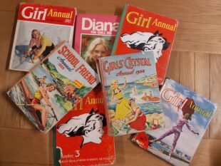 A selection of girls' annuals from the GWL Collection, on a wooden table. The annuals are from the 1950s, 60s and 70s, and have illustrations or photos of girls, some doing activities or sports. All the girls are white.