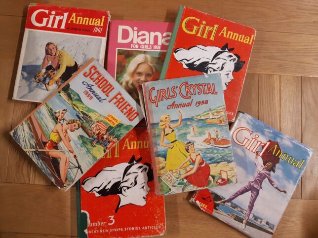 A selection of girls' annuals from the GWL Collection, on a wooden table. The annuals are from the 1950s, 60s and 70s, and have illustrations or photos of girls, some doing activities or sports. All the girls are white. 