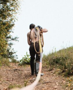 Artwork by Hannah Harkes: As Long As We Make It. A figure walks away from the camera along a bath, different braids of rope over their shoulder.